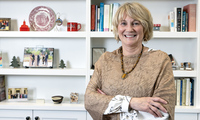 A smiling woman with short blonde hair stands in front of white bookshelves filled with books and decorative items, wearing a textured brown top, a necklace, and earrings, with her arms crossed.