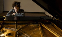 A person with curly hair leans on a grand piano, looking toward the camera. The pianos strings and internal components are visible in the foreground.