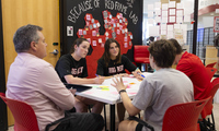 Four people sit around a small white table in discussion, with papers and markers on the table, in a classroom with red chairs and a wall.