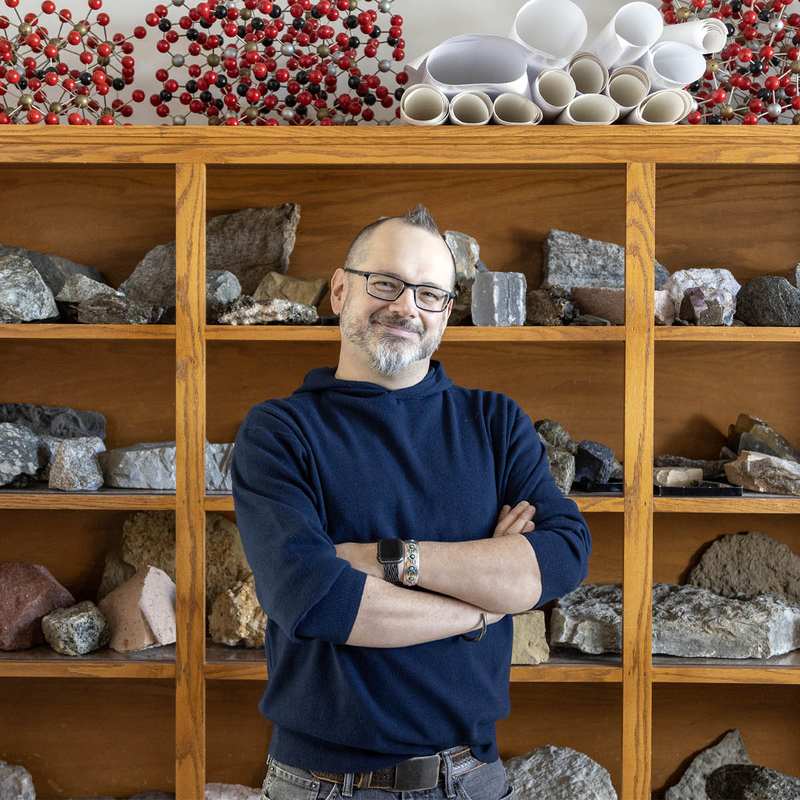 Photo of Geosciences Associate Professor Erik Klemetti standing in front of several wood shelves displaying various types of rocks