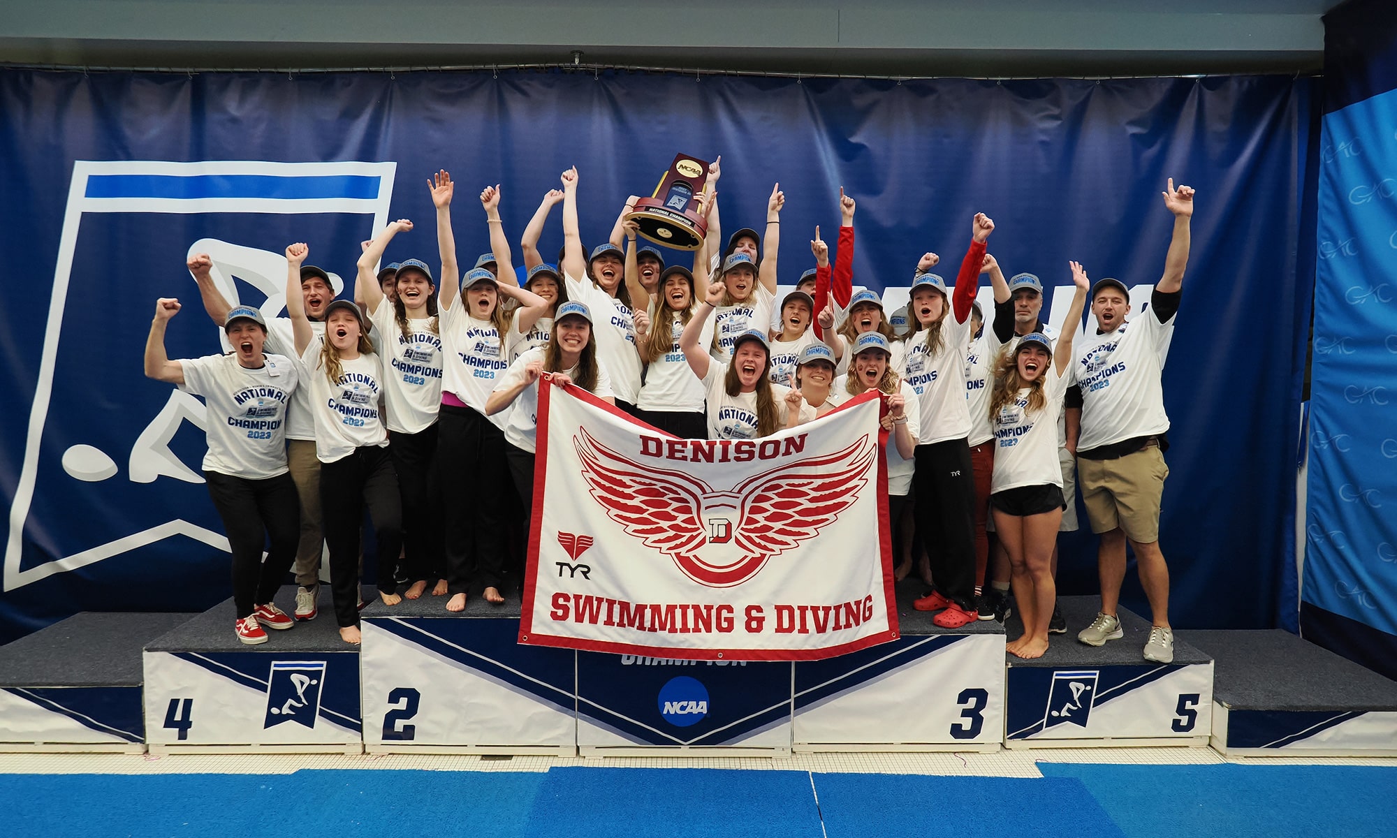 2023 Denison swim & dive team standing on the podium holding a trophy and a flag