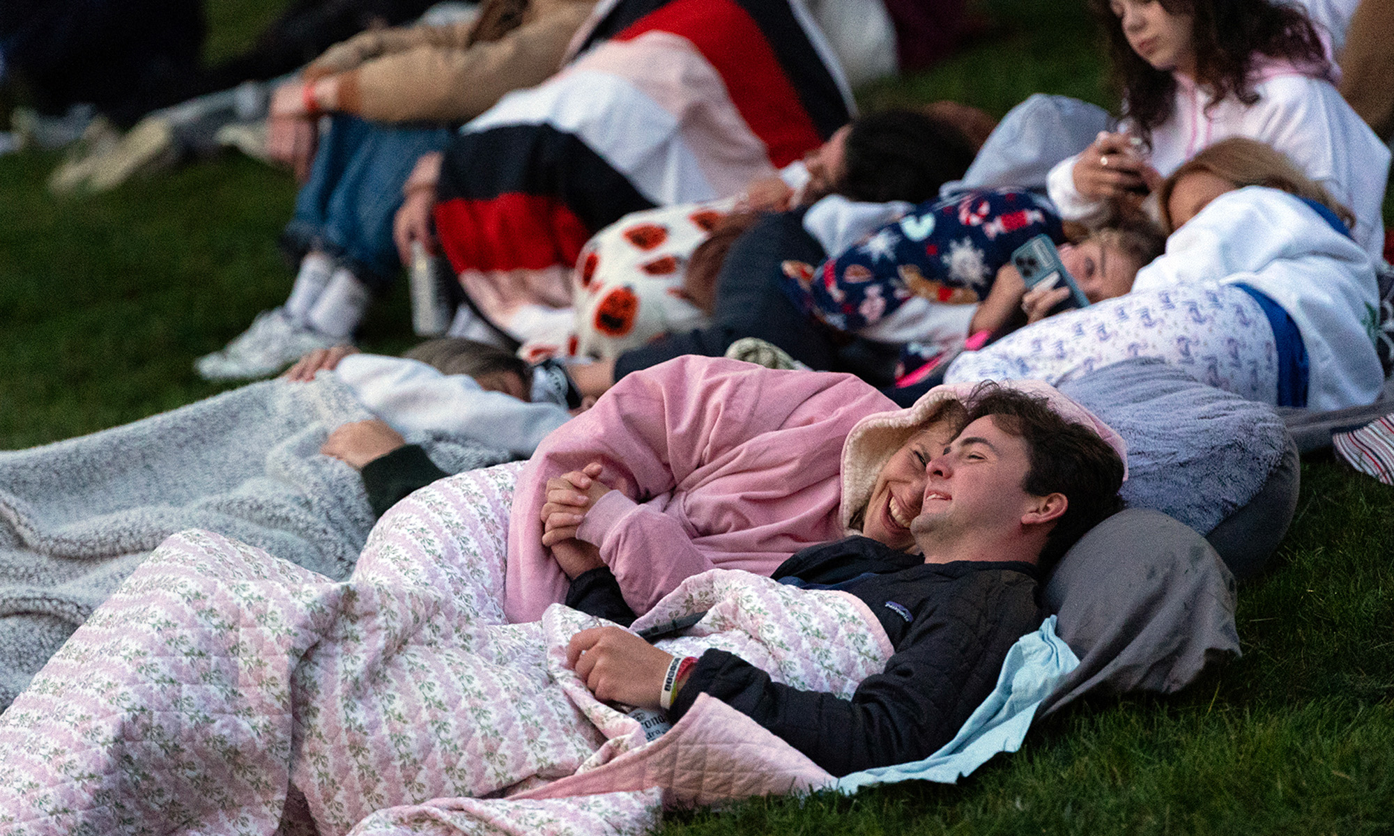 Two students cuddle in blankets on the ground, surrounded by other students awaiting a sunrise.