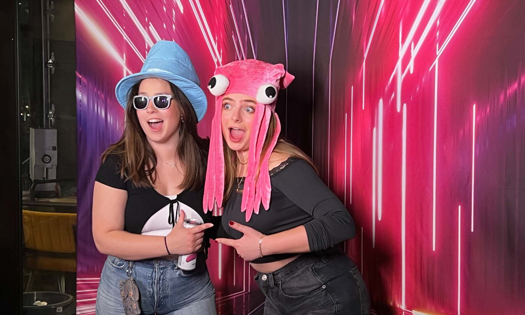 Two women in silly hats standing in front of a neon pink back drop 