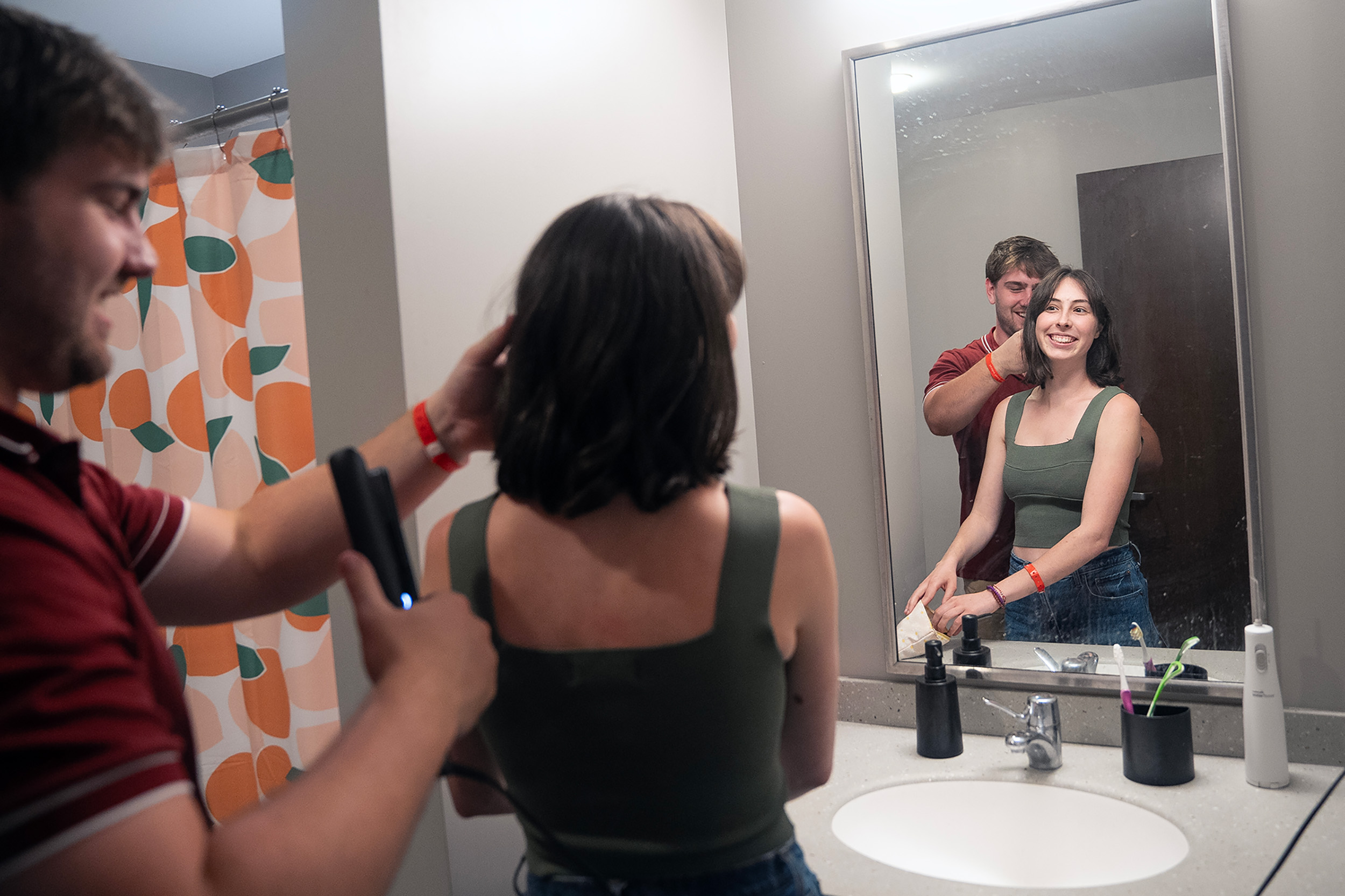 A man helps a woman straighten her hair as they both look into a bathroom mirror. 